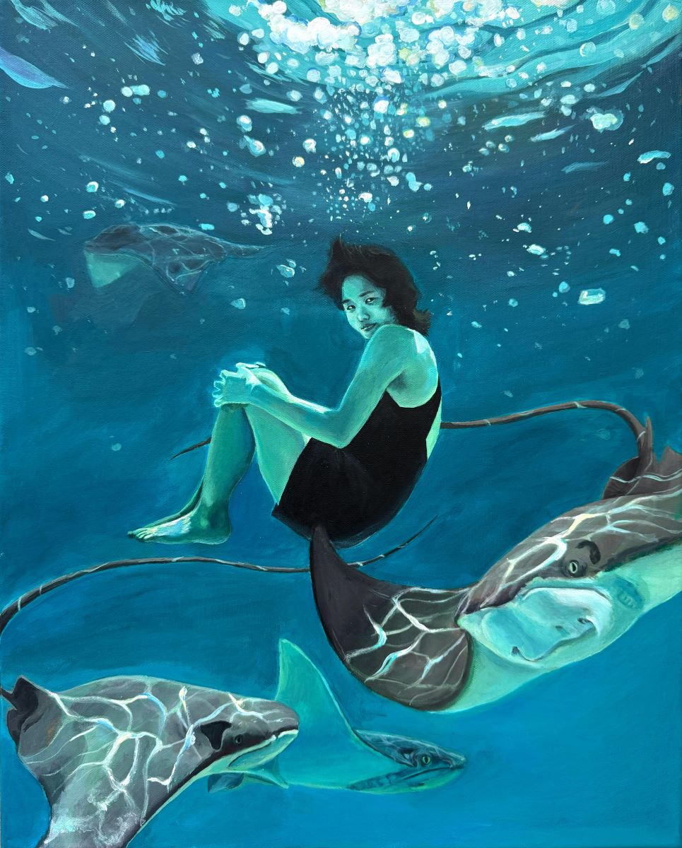A girl is under the water in the ocean with what appear to be stingrays 
							swimming underneath her. She wears a dark one-piece swim suit and holds her 
							knees as she looks to the viewer. Light is seen at the surface of the water.