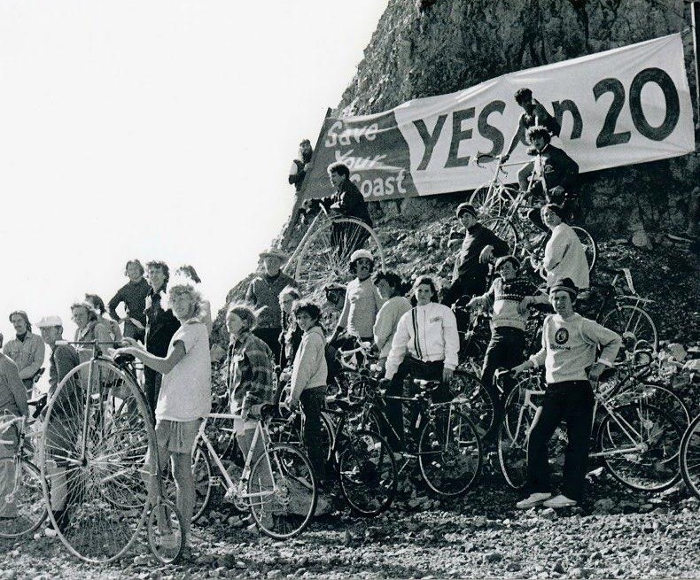 A black and white photo shows people with bikes standing next to a coastal bluff and a large Yes on 20 banner