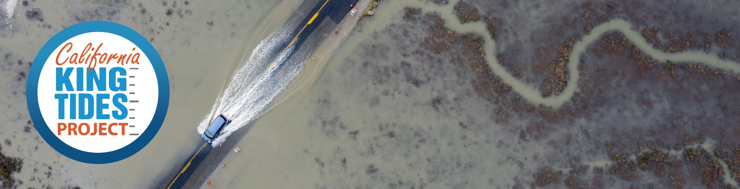 California King Tides Project. Seen from above, a car drives through a roadway flooded by the marshland on either side.