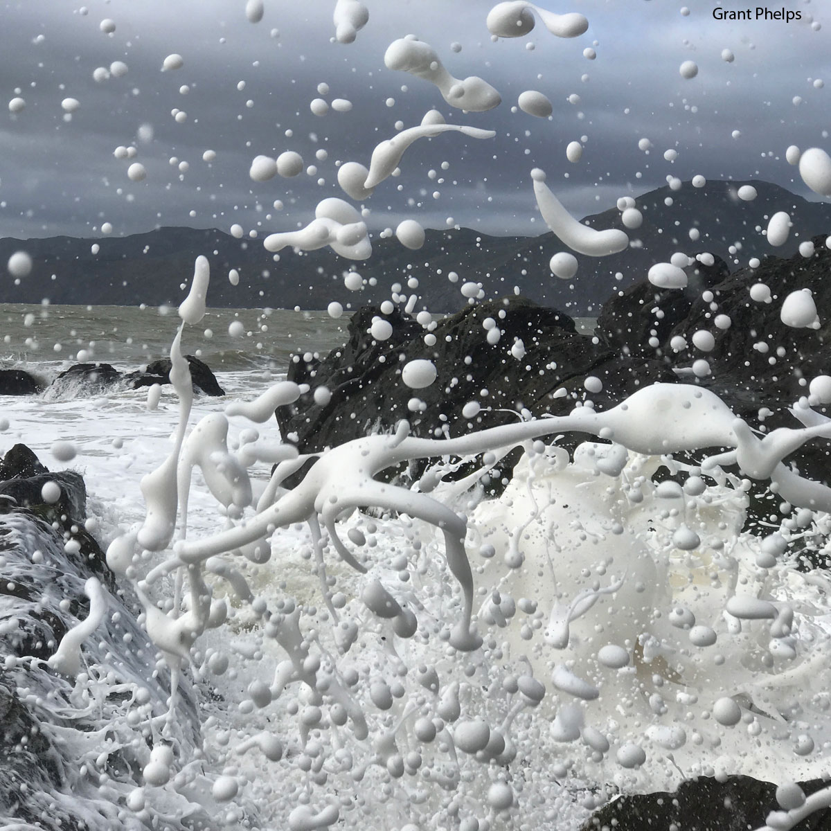 Photo of a gray ocean and sky behind dark rocks, dark hills in the background, white bubbles of foam directly in front of the camera