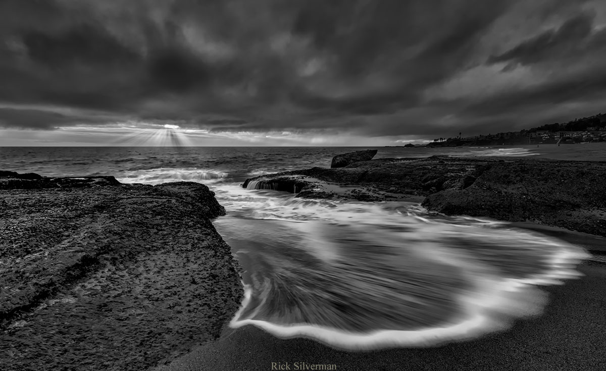 Photo of surf coming up onto the sand, light streaming through dark clouds, in black and white, by Rick Silverman