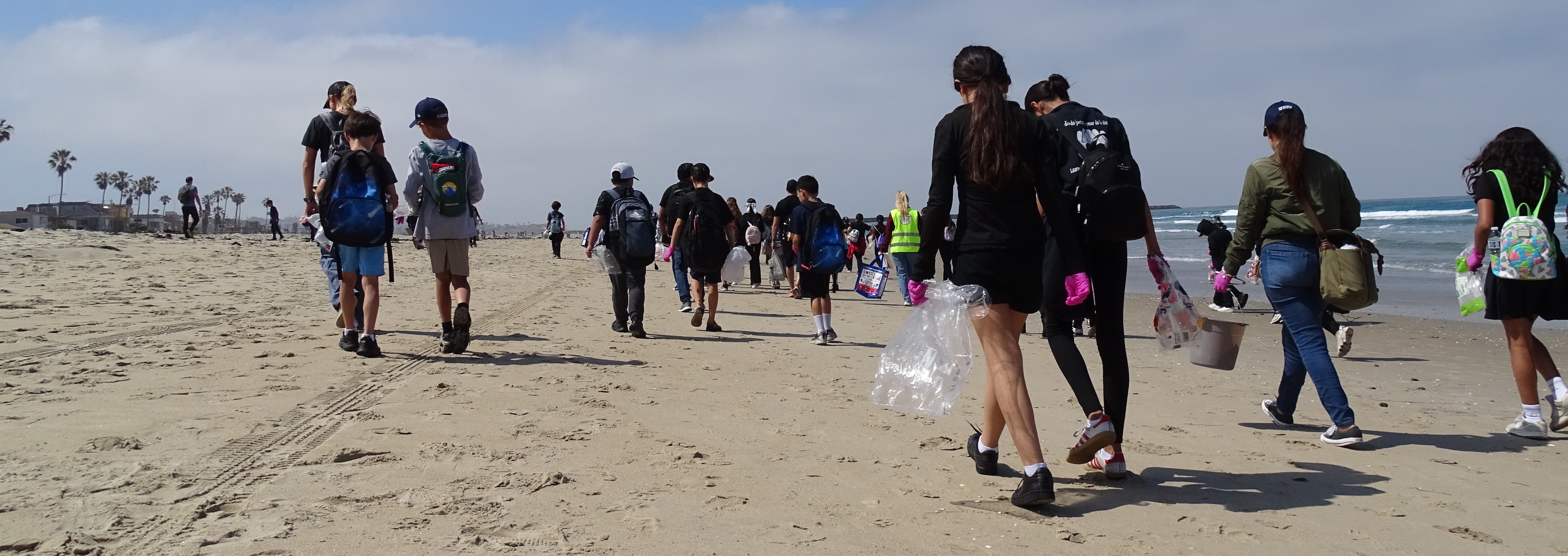 A large group of youth and adults walk along a beach carrying bags and buckets