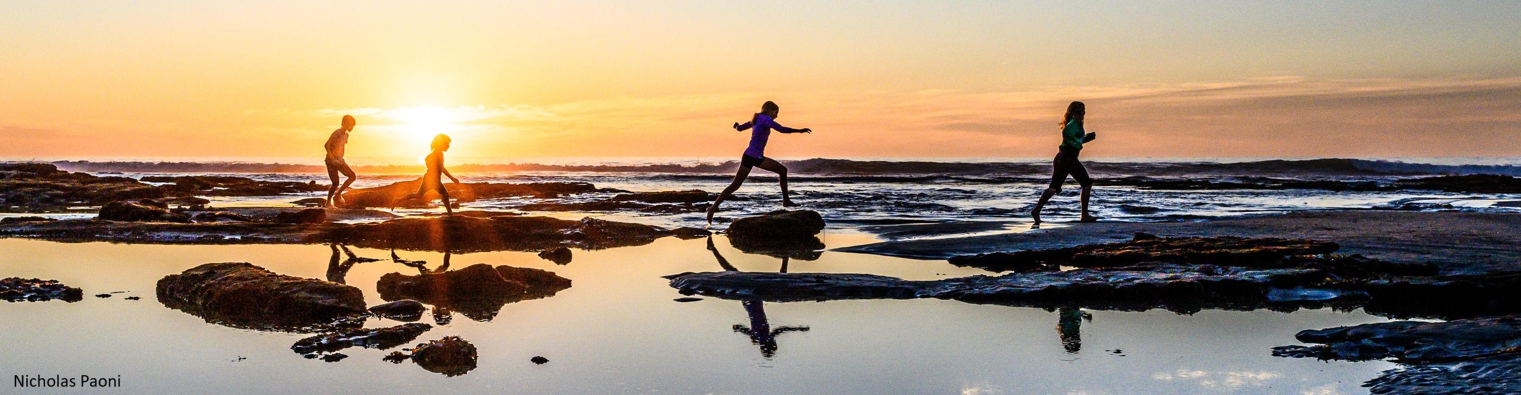 Children are running across a wet beach with the sunset behind them.
