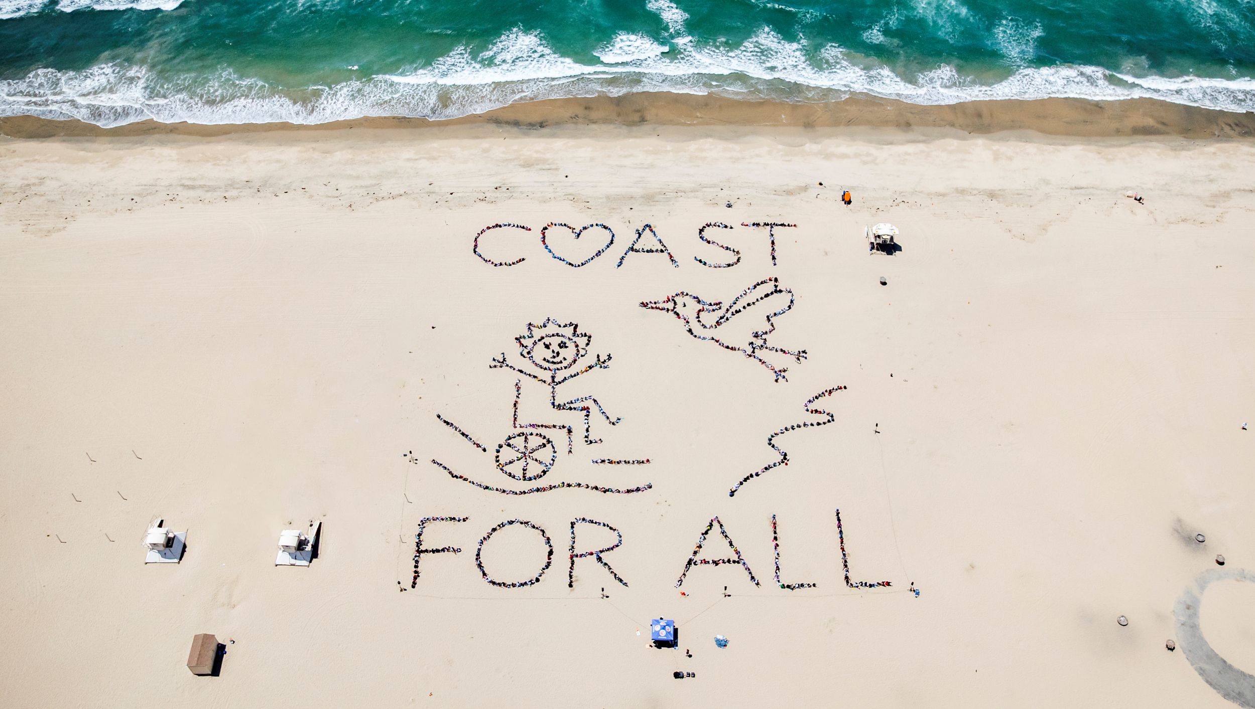 Orange County Kids' Ocean Day aerial artwork. People on the sand form an image of a person in a wheelchair, with a bird flying by, and the text, COAST FOR ALL, with the O in Coast in the shape of a heart.