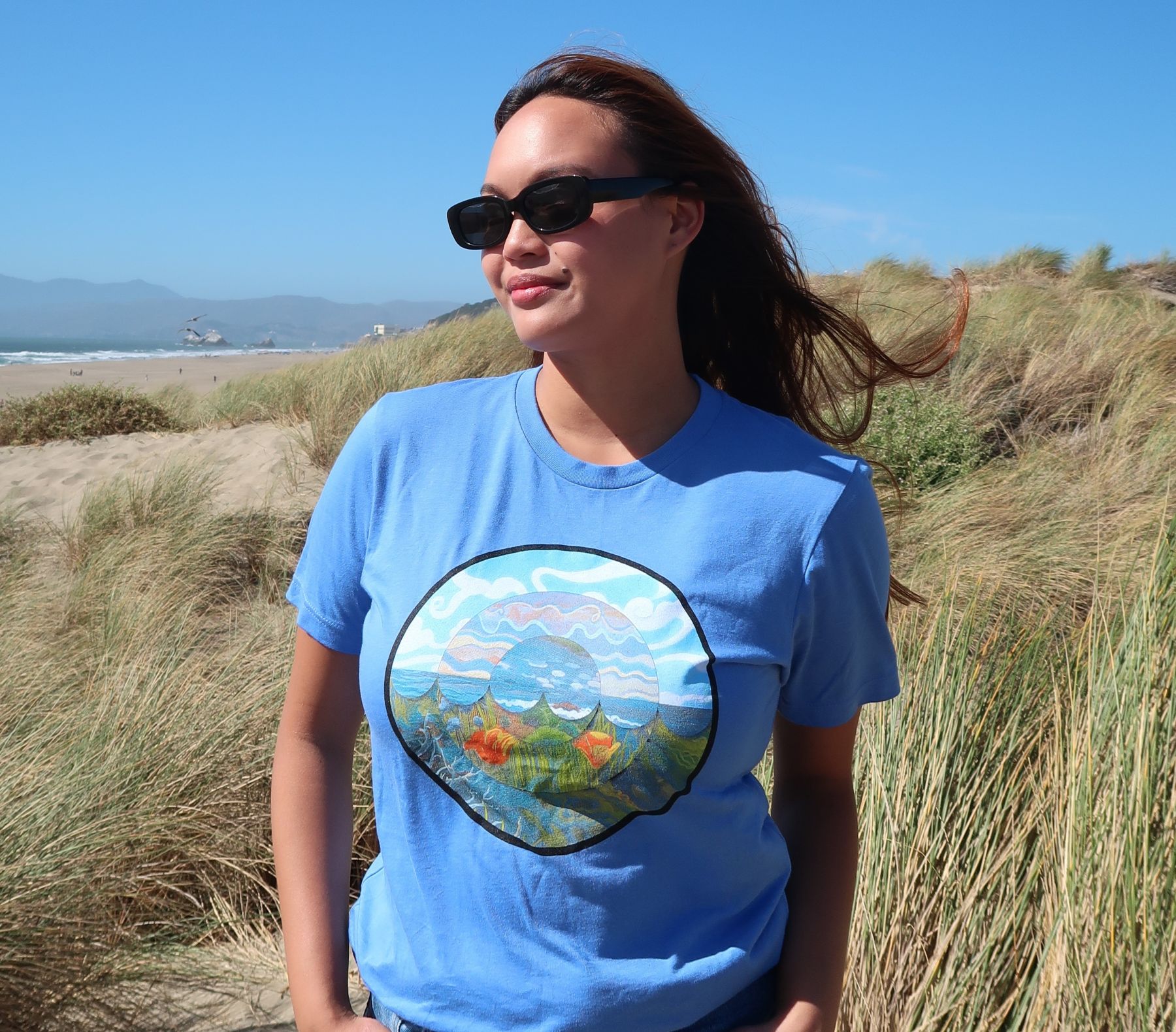 A woman stands in sand dunes wearing the 2026 
									Coastal Cleanup Day shirt. Shirt is light blue and has a round design on the front with the sky, ocean, and 
									California poppies.