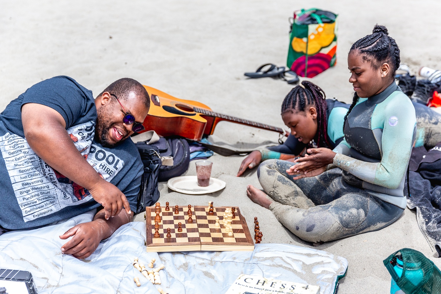 An adult and two youth sit and lounge on the sandy beach playing chess.
