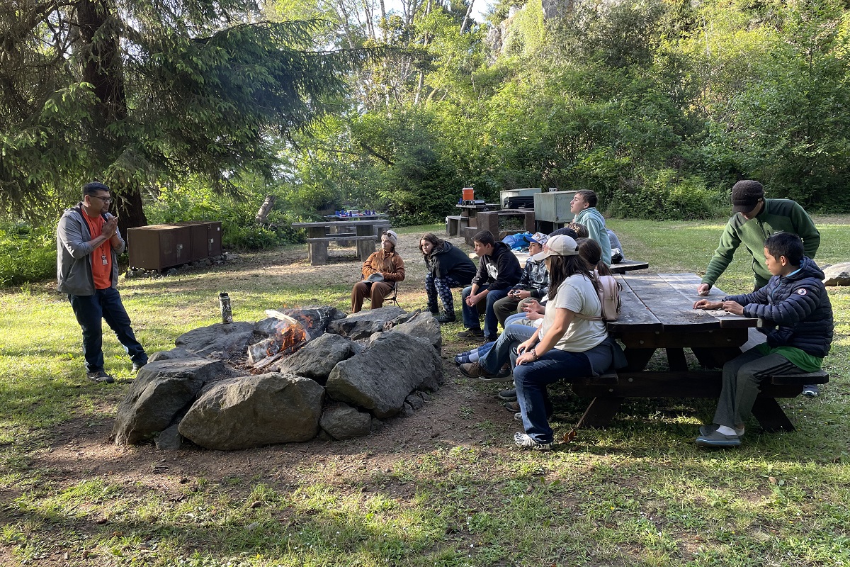 7th grade students sit at a picnic table next to a fire listening closely to a Yurok community member