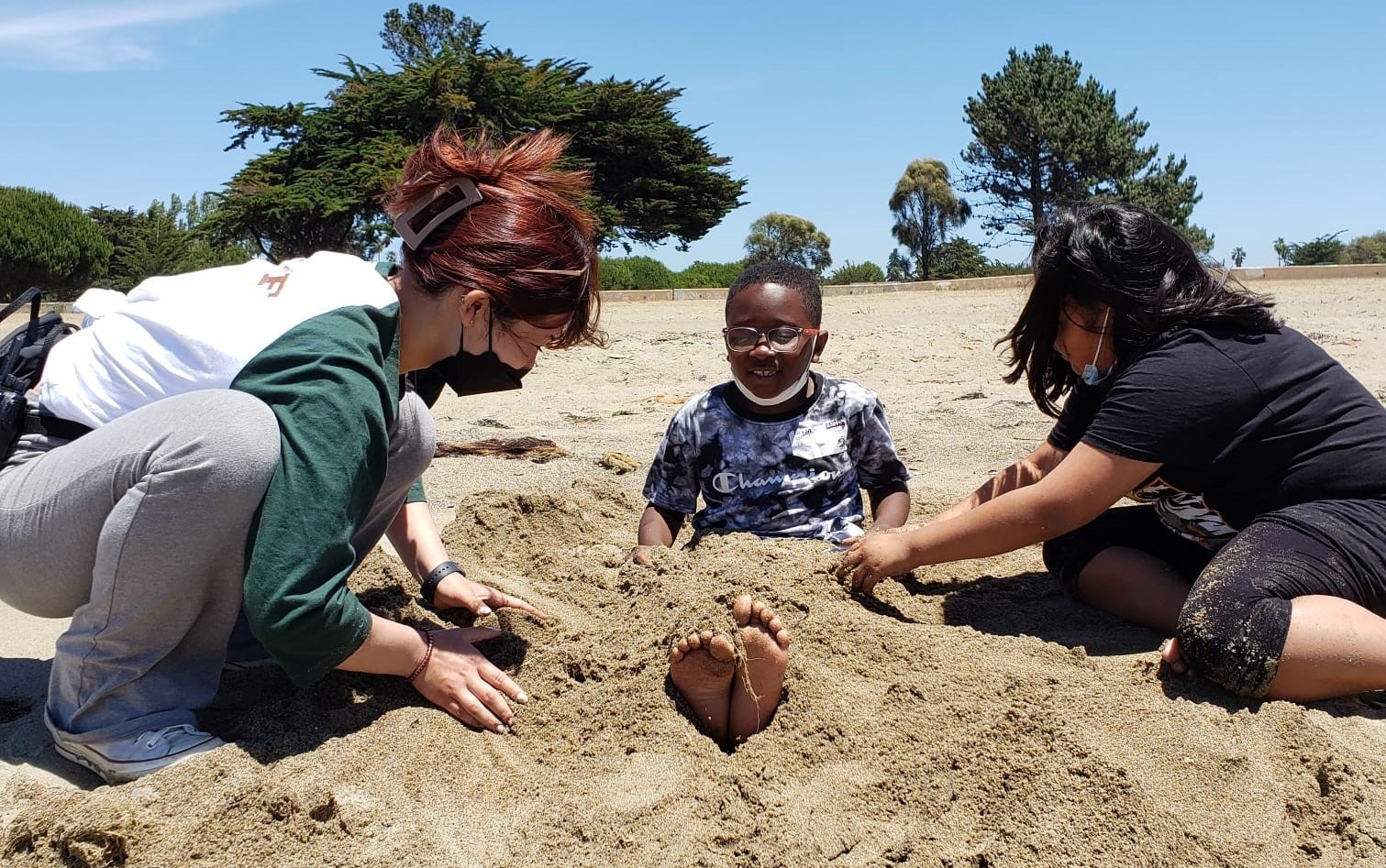 A young boy gets his legs buried in the sand by another child and an adult