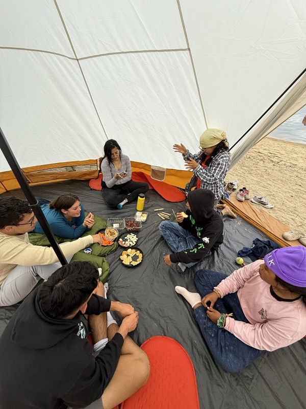 Seven young people sit in a circle inside a tent with snacks in the middle. One person has their hands out in mid-conversation. The open tent door
				reveals a sandy beach and coastal waters in the corner.