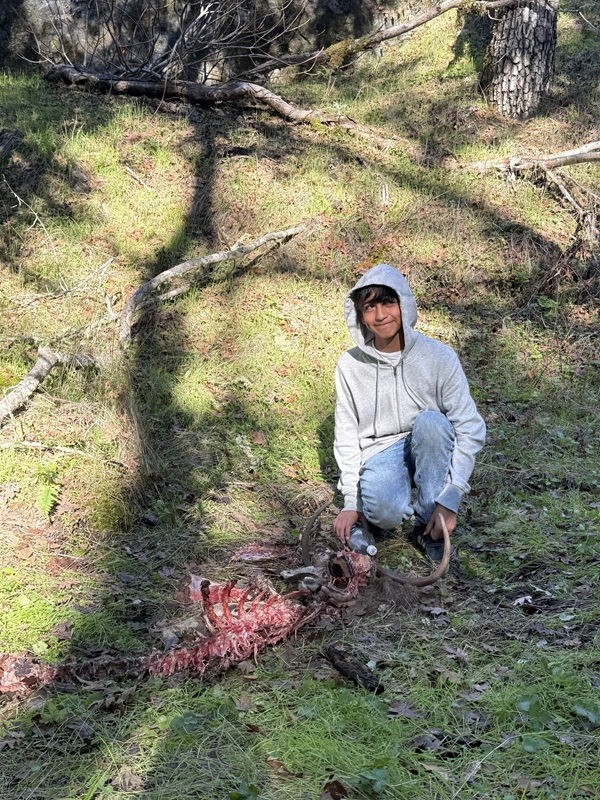 A youth with a sly smile and eyes looking to the side is crouched down next to deer remains. Green grass surrounds the youth and deer remains with 
				some tree trunks in the background.
