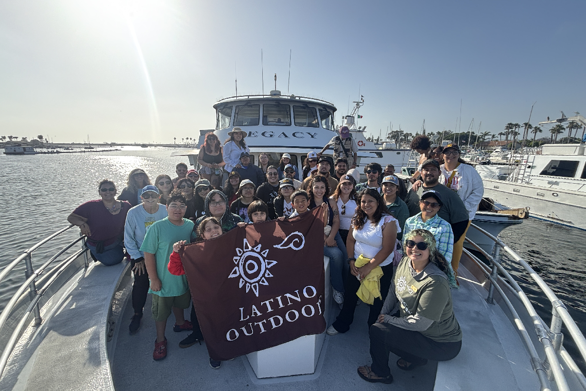 Over thirty whale watching participants of various ages smile on a boat with the sun and clear skies behind them. 
				Three children in front hold up a Latino Outdoors banner.