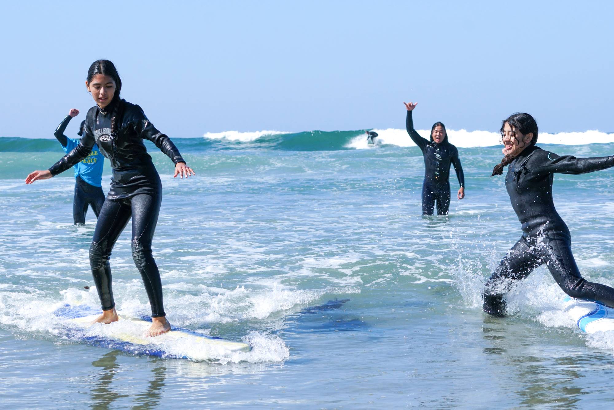 Two children succeed in standing on their boards in the surf while adults in the background cheer