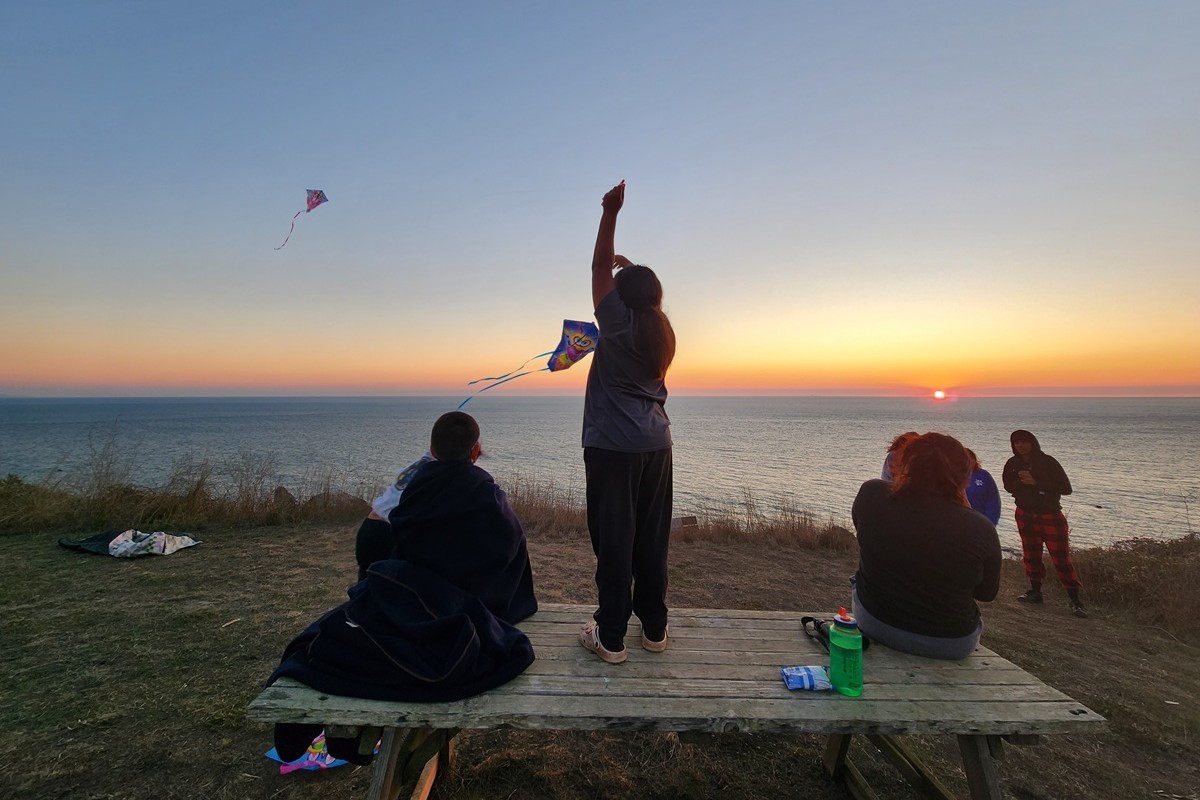 Three youth sit and stand on a picnic table flying kites. Another group of three youth stand on the grassy coastline
				in front of the sweeping ocean views. The sun is setting just visible over the water. The sky is a gradient from blue to orange.